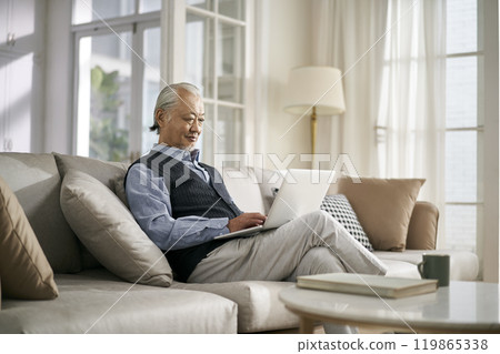 senior asian man sitting on couch looking at notebook computer senior asian man sitting on couch looking at notebook computer 119865338