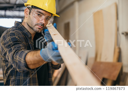 Construction worker in a yellow hardhat and safety glasses inspecting a piece of wood. Ideal for carpentry, woodworking, and quality control, highlighting craftsmanship in the workshop. labor day Construction worker in a yellow hardhat and safety glasses inspecting a piece of wood. Ideal for carpentry, woodworking, and quality control, highlighting craftsmanship in the workshop. labor day 119867873