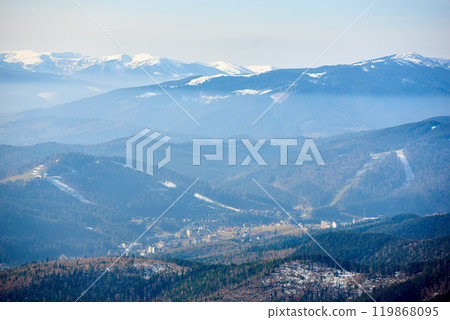 Aerial view of layered mountain ranges partially covered in snow. Valleys filled with dense forests and scattered settlements, while distant snow-capped peaks rise majestically under pale blue sky. Aerial view of layered mountain ranges partially covered in snow. Valleys filled with dense forests and scattered settlements, while distant snow-capped peaks rise majestically under pale blue sky. 119868095