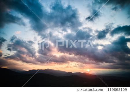 Aerial view breathtaking sunset over forest. Sun's rays break through dramatic clouds, casting golden glow across rolling hills and valleys. Distant mountains creating serene and picturesque scene. Aerial view breathtaking sunset over forest. Sun's rays break through dramatic clouds, casting golden glow across rolling hills and valleys. Distant mountains creating serene and picturesque scene. 119868109