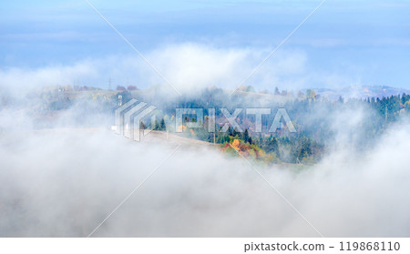 Picturesque view of hillside adorned with vibrant autumn foliage, ranging from rich greens to fiery reds and golden yellows. Wisps of fog drift across landscape under bright blue sky. 119868110