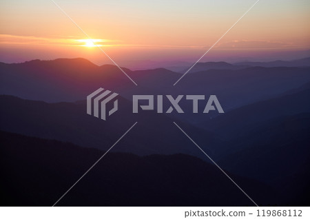 Sun sets over mountains, casting warm golden glow across sky. Layers of hills fade into soft shades of purple and blue. Peaceful ambiance of sunset highlights natural beauty of landscape. 119868112
