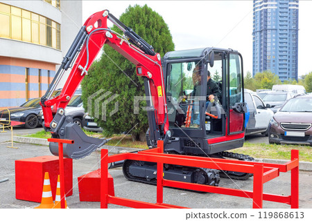 Compact red excavator at construction site with caution cones, operator visible, urban background, industrial machinery concept Compact red excavator at construction site with caution cones, operator visible, urban background, industrial machinery concept 119868613