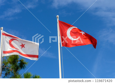 Turkish and Turkish Republic of Northern Cyprus flags waving under a clear blue sky during a sunny day at a coastal location 119871420