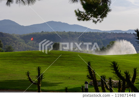 Serene golf course landscape with misty fountain and distant mountains under bright sunlight in a tranquil afternoon setting Serene golf course landscape with misty fountain and distant mountains under bright sunlight in a tranquil afternoon setting 119871448
