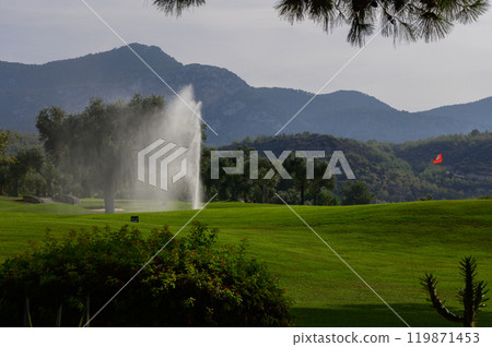 A serene landscape featuring a golf course with a water fountain amidst rolling hills under a clear sky in the early morning light 119871453