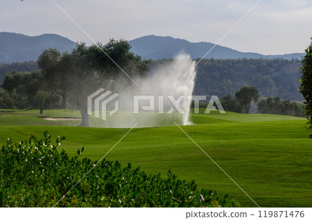 A serene morning at a lush green golf course with a gentle fountain misting under the sun and distant mountains as a backdrop A serene morning at a lush green golf course with a gentle fountain misting under the sun and distant mountains as a backdrop 119871476
