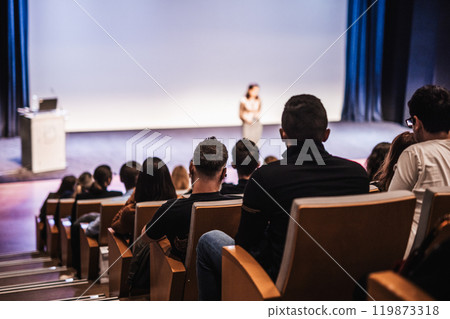 Business and entrepreneurship symposium. Female speaker giving a talk at business meeting. Audience in conference hall. Rear view of unrecognized participant in audience. 119873318