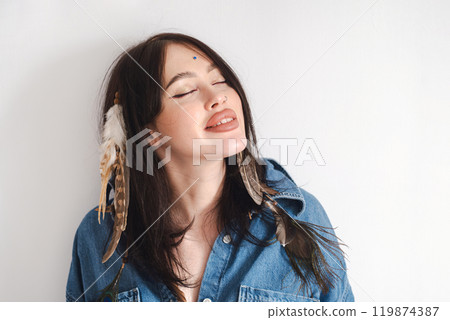 Smiling woman with feathers in hair wearing denim shirt. Embodying a free-spirited vibe. 119874387