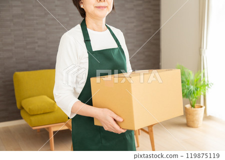 A senior female housewife/housekeeper wearing a green apron holding a cardboard box in the living room at home A senior female housewife/housekeeper wearing a green apron holding a cardboard box in the living room at home 119875119