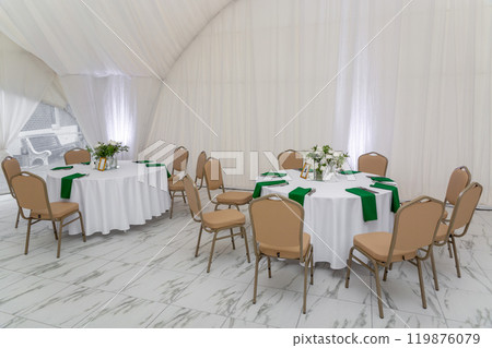 Wine glasses, silver tableware and green napkins on round tables covered with white tablecloth. 119876079