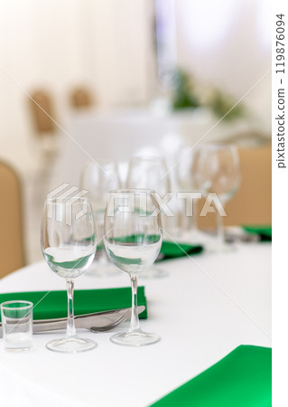 Close up of wine glasses, silver tableware and green napkins on round tables covered with white tablecloth. 119876094