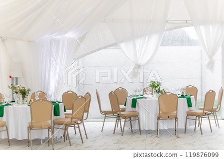 Wine glasses, silver tableware and green napkins on round tables covered with white tablecloth. 119876099