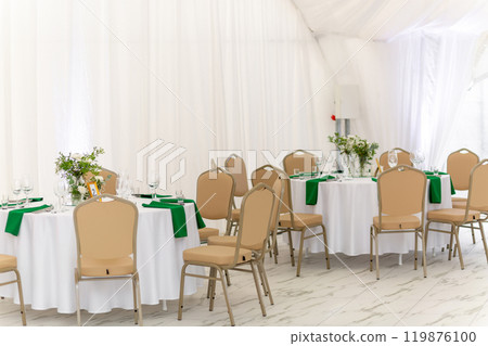 Wine glasses, silver tableware and green napkins on round tables covered with white tablecloth. Wine glasses, silver tableware and green napkins on round tables covered with white tablecloth. 119876100