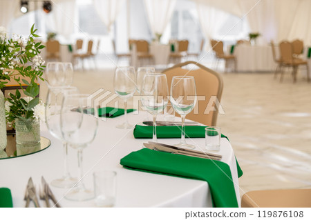 Close up of wine glasses, silver tableware and green napkins on round tables covered with white tablecloth. Close up of wine glasses, silver tableware and green napkins on round tables covered with white tablecloth. 119876108