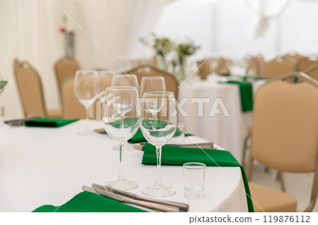 Close up of wine glasses, silver tableware and green napkins on round tables covered with white tablecloth. 119876112