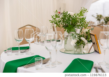 Close up of wine glasses, silver tableware and green napkins on round tables covered with white tablecloth. 119876113