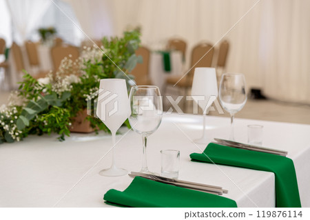 Close up of wine glasses, silver tableware and green napkins on round tables covered with white tablecloth. 119876114