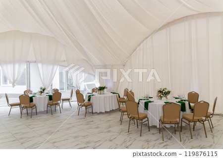 Wine glasses, silver tableware and green napkins on round tables covered with white tablecloth. 119876115