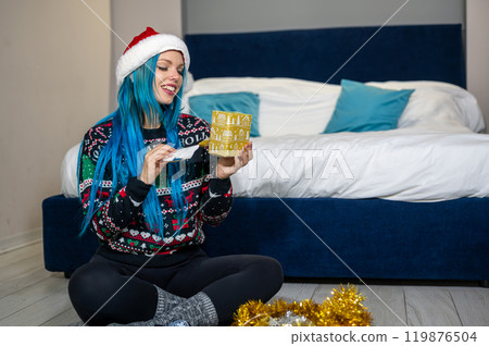 Portrait of a beautiful caucasian hipster woman in Christmas clothes. She is sitting next to the bed and smilingly unwrapping a package. Expectation, surprise, happiness. Portrait of a beautiful caucasian hipster woman in Christmas clothes. She is sitting next to the bed and smilingly unwrapping a package. Expectation, surprise, happiness. 119876504