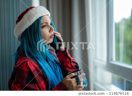 A beautiful hipster caucasian woman in Christmas clothes. She is talking on her cell phone while looking out the window, she has a cup of tea. Christmas,talking with friends, positivity and happiness. A beautiful hipster caucasian woman in Christmas clothes. She is talking on her cell phone while looking out the window, she has a cup of tea. Christmas,talking with friends, positivity and happiness. 119876516