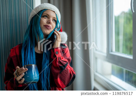 A beautiful hipster caucasian woman in Christmas clothes. She is talking on her cell phone while looking out the window, she has a cup of tea. Christmas,talking with friends, positivity and happiness. A beautiful hipster caucasian woman in Christmas clothes. She is talking on her cell phone while looking out the window, she has a cup of tea. Christmas,talking with friends, positivity and happiness. 119876517