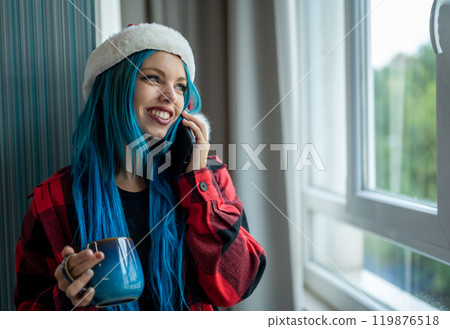 A beautiful hipster caucasian woman in Christmas clothes. She is talking on her cell phone while looking out the window, she has a cup of tea. Christmas,talking with friends, positivity and happiness. 119876518