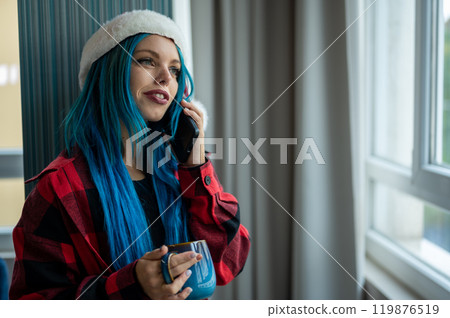A beautiful hipster caucasian woman in Christmas clothes. She is talking on her cell phone while looking out the window, she has a cup of tea. Christmas,talking with friends, positivity and happiness. 119876519