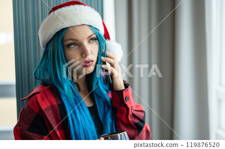 A beautiful hipster caucasian woman in Christmas clothes. She is talking on her cell phone while looking out the window, she has a cup of tea. Christmas,talking with friends, positivity and happiness. 119876520