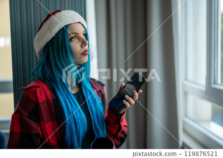 A beautiful hipster caucasian woman in Christmas clothes. She is talking on her cell phone while looking out the window, she has a cup of tea. Christmas,talking with friends, positivity and happiness. 119876521