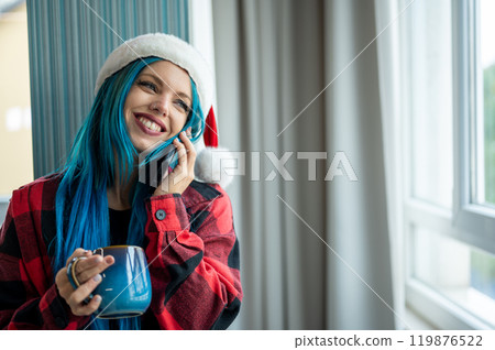 A beautiful hipster caucasian woman in Christmas clothes. She is talking on her cell phone while looking out the window, she has a cup of tea. Christmas,talking with friends, positivity and happiness. 119876522