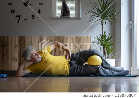 Elderly man in bright yellow tshirt practicing pilates exercise with a ball 119876807