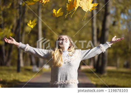 Beautiful young woman in an autumn park scatters maple leaves. 119878103
