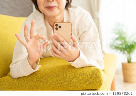 A middle-aged to senior woman lying on the sofa and making a circle sign while looking at her smartphone 119878261