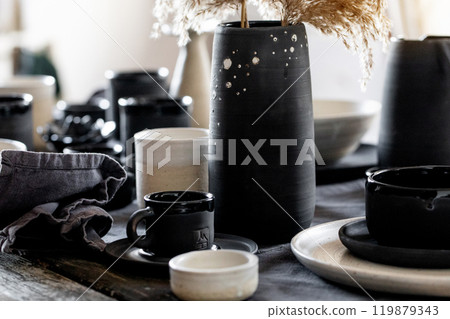 Rustic table setting with empty craft handmade ceramic tableware, black and white rough bowls, cups, jug and vase on linen tablecloth. Dry reed flowers. Day light dining room. 119879343