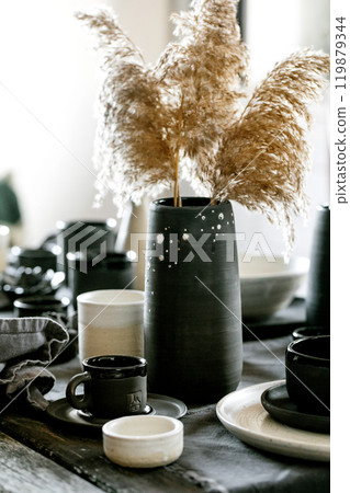 Rustic table setting with empty craft handmade ceramic tableware, black and white rough bowls, cups, jug and vase on linen tablecloth. Dry reed flowers. Day light dining room. 119879344