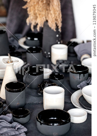 Rustic table setting with empty craft handmade ceramic tableware, black and white rough bowls, cups, jug and vase on linen tablecloth. Dry reed flowers. Day light dining room. 119879345