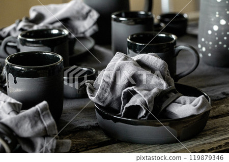 Rustic table setting with empty craft handmade ceramic tableware, black bowls, plates, cups, jug and vase on linen tablecloth. Blck ceramic roses flowers decor. Day light dining room. 119879346