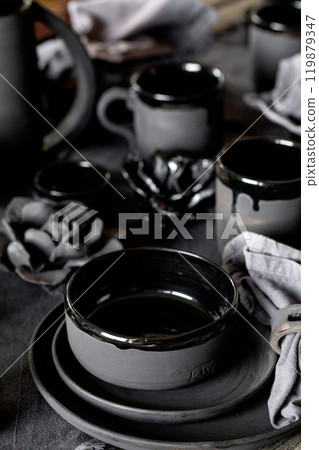 Rustic table setting with empty craft handmade ceramic tableware, black bowls, plates, cups, jug and vase on linen tablecloth. Blck ceramic roses flowers decor. Day light dining room. 119879347