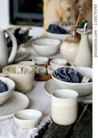Rustic table setting with empty craft handmade ceramic tableware, white rough bowls, plates, cups, jug and vase on linen tablecloth. Dry reed flowers. Day light dining room. 119879349