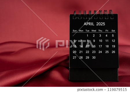 Black desk calendar for April 2025 on a red satin or silk textile background. 119879515