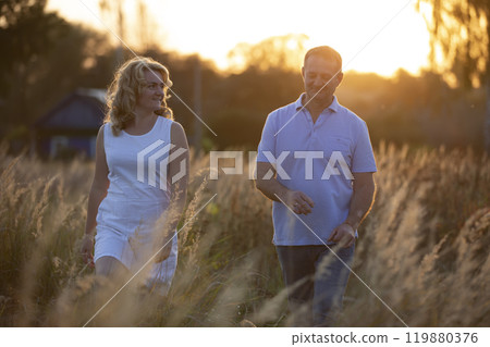 A beautiful middle-aged couple in love are walking on the field against the background of sunset. A beautiful middle-aged couple in love are walking on the field against the background of sunset. 119880376