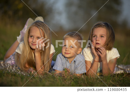 Sisters and little brother lie on the grass. Children play in the meadow. A happy family. 119880435
