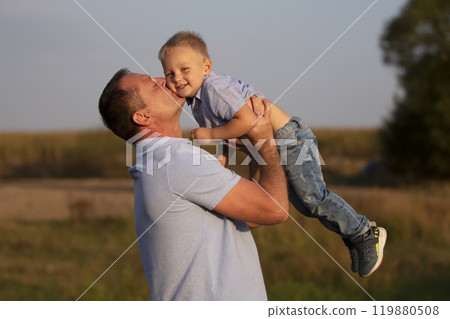 Happy dad plays with his son on the background of the field. Joyful grandfather kisses his grandson. Happy dad plays with his son on the background of the field. Joyful grandfather kisses his grandson. 119880508