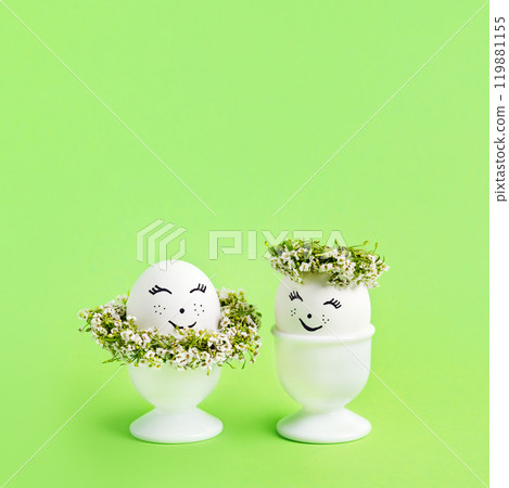 Two white Easter eggs with funny face with spring flowers in white stands on green background. Happy Easter concept. Greeting card. 119881155