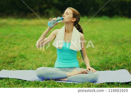 Fitness woman drinking water from bottle on a mat on the grass in summer park 119881920