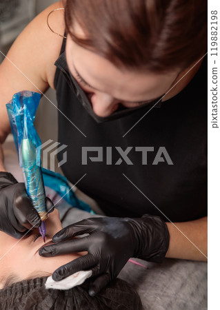 Cosmetologist applying permanent makeup on eyebrows with tattoo machine Cosmetologist applying permanent makeup on eyebrows with tattoo machine 119882198