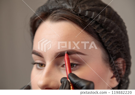 A cosmetologist draws the shape of eyebrows on a young woman's face with a contouring pencil 119882220