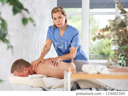 Female masseur performs back stroking and patting during massage session for male client Female masseur performs back stroking and patting during massage session for male client 119882304