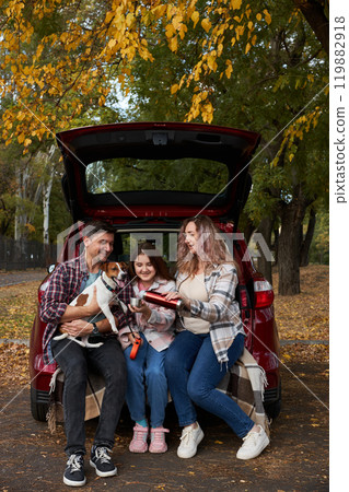 Happy parents with daughter and cute dog Jack Russel terrier sitting in car trunk on autumn day 119882918